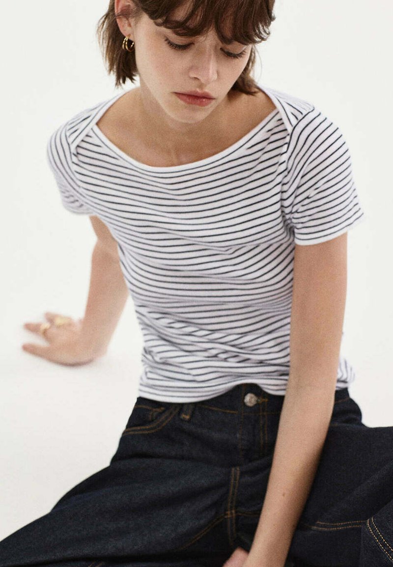 Jeune femme assise, portant un t-shirt à manches courtes rayé blanc et noir et un jean en denim foncé, regardant vers le bas.