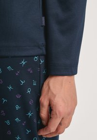 Marineblauwe longsleeve top met een zachte textuur en een stoffen label; gecombineerd met donkere, gemusterde broeken met kleurrijke astrologische symbolen.