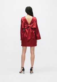 Kvinne med flettehår iført en rød satengkåpe med stor sløyfe på ryggen og svarte høye hæler.