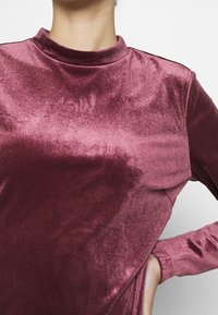 Bordový sametový top s dlouhým rukávem, vysokým límcem a přiléhavým střihem. Hladký povrch se subtilním leskem, s zakřiveným lemem a úzkými manžetami.