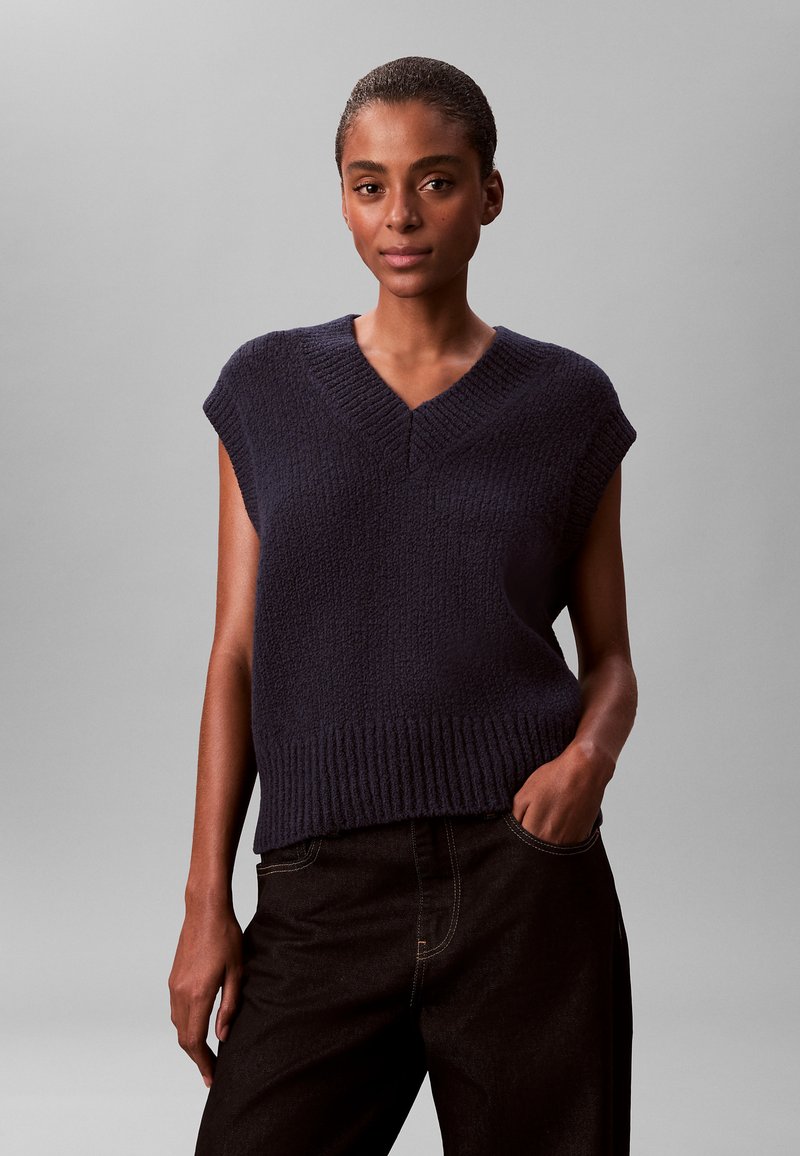 Mujer con cabello corto que lleva un chaleco de punto sin mangas color azul marino oscuro y pantalones negros, de pie con una mano en el bolsillo frente a un fondo gris.