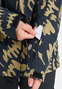 Tarnmuster-Jacke in Dunkelblau und Beige, mit einer Reißverschlusstasche am Arm. Verfügt über einen Velcro-Bund zur Anpassung der Passform und zum Wetterschutz.