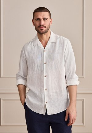 Homme aux cheveux courts portant une chemise en lin blanche boutonnée avec manches retroussées et un pantalon foncé, debout contre un mur beige à panneaux.