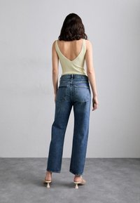 Camiseta sin mangas verde claro con textura acanalada y escote en V profundo en la espalda, combinada con jeans azules de cintura alta y tacones beige con un diseño único de tacón esculpido.