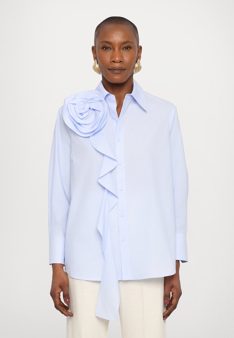 Ανοιχτό μπλε πουκάμισο με φουσκωτό μπροστινό μέρος και μεγάλο λουλούδι από ύφασμα στον αριστερό ώμο. Μακριά μανίκια, προσαρμοσμένη εφαρμογή, λεία υφή.