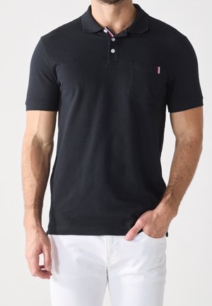 Mann trägt schwarzes Kurzarm-Poloshirt mit Brusttasche und weiße Hose, Hand in der Tasche, vor hellem einfarbigem Hintergrund.
