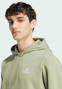 Olivgrön hoodie i mjukt material med en snörning i huvan. Har en vit Adidas-logotyp på bröstet. Slät textur, avslappnad passform.