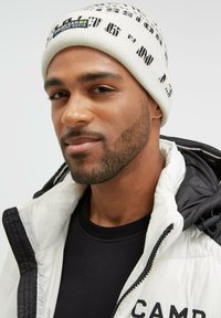 White knit beanie with black pattern and label, paired with a black jacket featuring a puffy white collar. Focus on materials and textures.