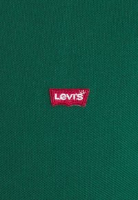 Tissu vert texturé avec une étiquette logo Levi's rouge, présentant une forme distinctive de batwing. Vue rapprochée mettant en avant le matériau et le branding.
