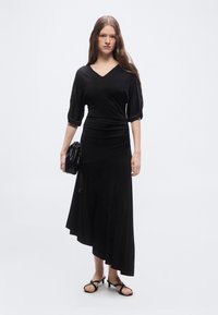 Γυναίκα με μαύρο ασύμμετρο μίντι φόρεμα με μανίκια μέχρι τον αγκώνα, κρατώντας μαύρο τσαντάκι με κορδόνι και φορώντας σανδάλια με χαμηλό τακούνι.