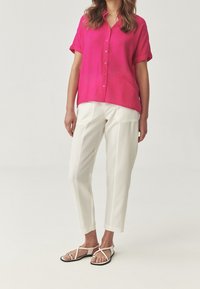 Camicia fucsia con bottoni e maniche corte, abbinata a pantaloni bianchi a gamba stretta. Sandali bianchi con cinturini e dettaglio argentato completano il look.