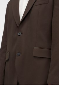 Gros plan d'un blazer marron avec deux boutons noirs, poches à rabat et une chemise blanche visible en dessous.