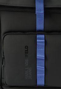 Μπροστινή τσέπη μαύρου σακιδίου με κάθετη μπλε λωρίδα και διακριτικό λογότυπο "KARL LAGERFELD JEANS" σε σκούρο χρώμα.