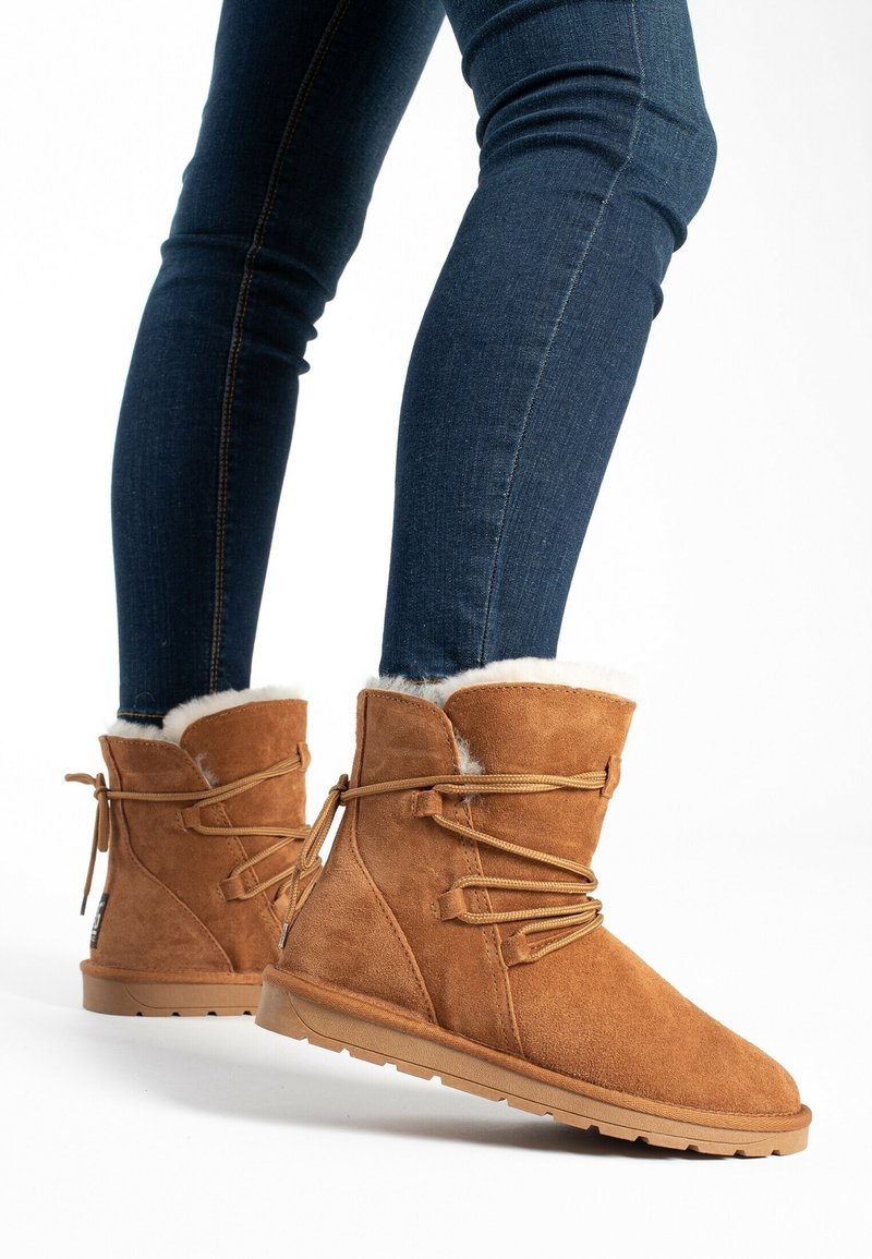 Beige Wildleder-Stiefeletten mit einem weichen weißen Futter, Schnürdetail und einer strukturierten Gummisohle, getragen mit dunkelblauen Jeans.