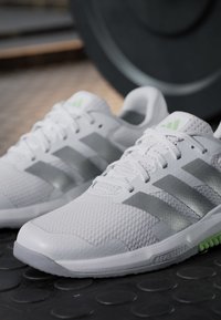 Chaussures de sport blanches avec des rayures grises et des lacets reposant sur un sol de gym noir texturé près d'une plaque de poids.