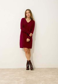 Burgundské pletené šaty s výstřihem do V a dlouhými rukávy, doplněné lesklými kotníkovými botami. Měkká textura, design po kolena.