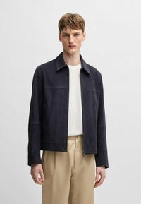 Navy suede jacket with a collared neckline, zip closure, and two side pockets. Features a fitted cut and smooth texture. Worn over a white shirt.