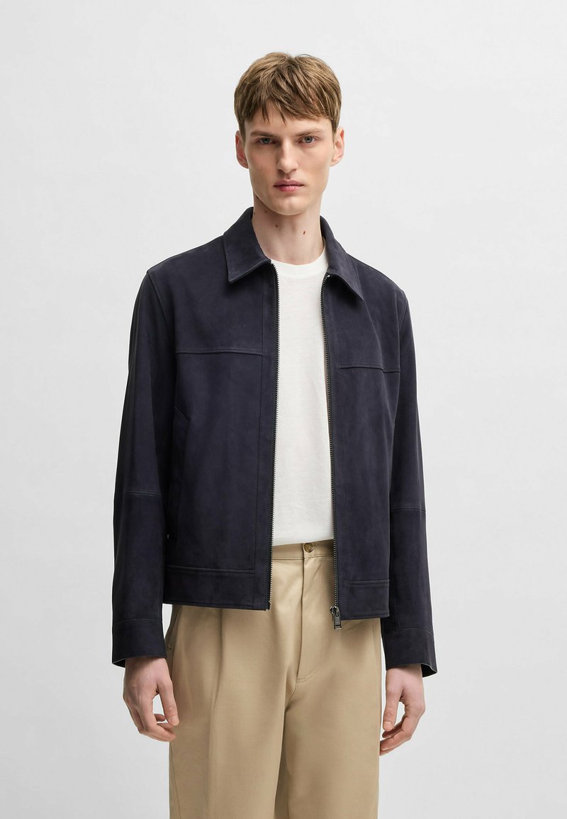 Navy suede jacket with a collared neckline, zip closure, and two side pockets. Features a fitted cut and smooth texture. Worn over a white shirt.