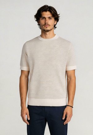 Giovane uomo con capelli corti e scuri indossa un maglione beige a maniche corte con texture e pantaloni scuri, in piedi davanti a uno sfondo chiaro e uniforme.