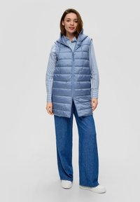 Helles blaues gepolstertes Weste mit Kapuze, ausgestattet mit einem Reißverschluss vorne und horizontalen Nähten. Tragen über einem gestreiften Hemd und weit geschnittenen blauen Hosen.