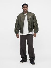 Homme debout portant une veste bomber vert olive, un t-shirt blanc, un pantalon cargo ample foncé et des baskets noires sur un fond blanc uni.