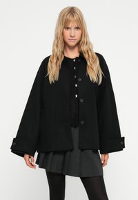 Svart oversized jacka med rund krage, stor knäppning och sidfickor. Har vida ärmar och en mjuk textur.