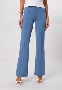 Pantalons évasés bleus en tissu extensible, avec des coutures accentuées, une taille haute et une texture lisse. Associés à des sandales blanches.