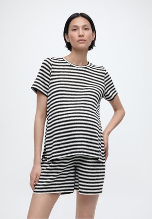 Lühikeste tumedate juustega naine, kes kannab musta ja valget horisontaaltriibulist lühikeste varrukatega toppi ja sobivaid lühikesi pükse, seisab ühetaolise heleda taustaga.