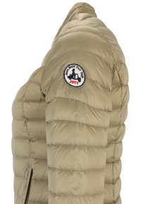 Beige pufferjakke med quiltet design, sidelukning og et rundt mærke på skulderen med tekst og en illustration.