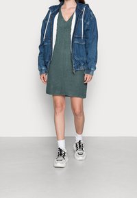 Veste en jean avec capuche portée sur une robe en maille verte. Baskets blanches avec accents noirs et chaussettes montant jusqu'à la cheville. Col montant et coupe ajustée.