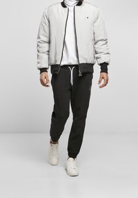 Man die loopt en witte sneakers, zwarte joggerbroek, witte coltrui en lichtgrijze bomberjack met zwarte bies draagt.