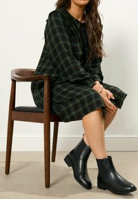 Vihreä ja musta ruudullinen mekko, jossa on napitettava etuosa, yhdistettynä mustiin nahkasaappaisiin nilkkoihin. Malli istuu puupenkillä.