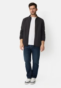 Dunkelgraue Jacke mit Reißverschluss und hohem Kragen, strukturiertem Stoff, Fronttasche, kombiniert mit einem weißen T-Shirt und blauen Jeans, dazu weiße Turnschuhe.