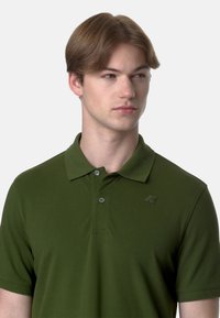 Polo shirt verde oliva in cotone misto, con patta a due bottoni, colletto piatto e un sottile logo sul petto. Texture liscia.