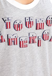 Maglietta manica senza con scollo rotondo bianco che presenta la scritta "YOUNG AMERICA" in caratteri bold. Le lettere hanno un motivo a strisce rosse e bianche con accenti blu.