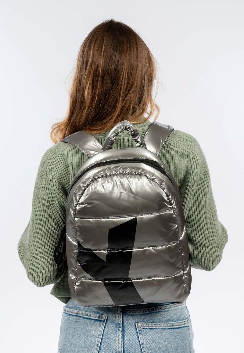 Personne aux longs cheveux bruns porte un pull en maille vert et un jean bleu clair, portant un sac à dos matelassé argenté brillant avec un design géométrique noir.
