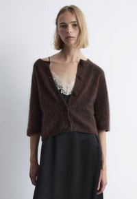 Braune, flauschige Strickjacke mit drei Knöpfen, 3/4- Ärmeln, die über einem schwarzen Kleid mit Spitze getragen wird. Weiche Textur und cropped Design.