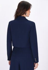 Woman with dark brown hair wears navy blue cropped blazer and matching trousers, showing back view against plain white background.