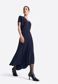 Granatowa sukienka z dekoltem w kształcie litery V i krótkimi rękawami, z falującym asymetrycznym dołem. Modelka nosi czarne mulesy na obcasie i przezroczyste rajstopy.