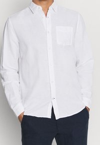 Camicia di lino bianca con bottoni e taschino sul petto, con colletto classico e maniche lunghe, abbinata a pantaloni scuri.