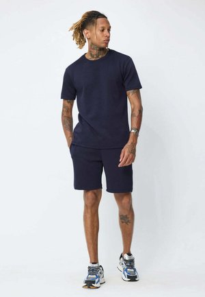 Homme avec des tatouages portant un t-shirt bleu marine et un short avec des baskets bleues et blanches, debout devant un fond uni.