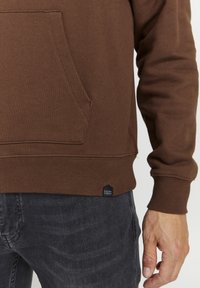 Brun sweatshirt med en kængurulomme, ribbede manchetter og kant. Har et lille sort mærke i bunden. Virker blød og struktureret.