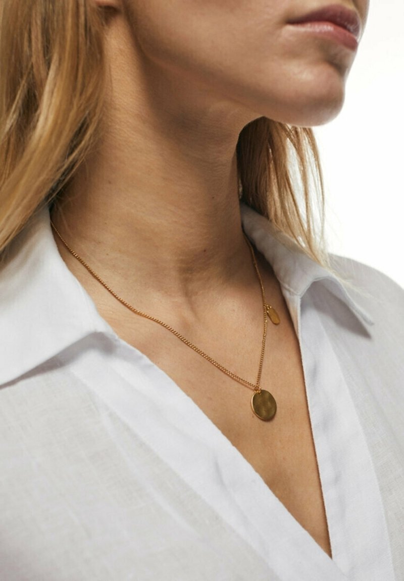 Gros plan sur le cou d'une femme portant une chemise blanche à col et une chaîne en or avec un pendentif rond.