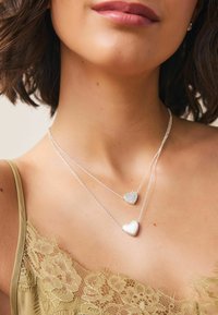 Deux colliers en argent avec des pendentifs en forme de cœur; l'un est lisse et blanc, l'autre est orné de petites pierres transparentes. Haut en dentelle de couleur verte olive.