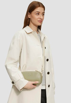 Hellgrüne Leder-Crossbody-Tasche mit einem Reißverschluss und einem Riemen, in einer Hand gehalten. Getragen mit einem beigen Trenchcoat über einem grauen Oberteil.