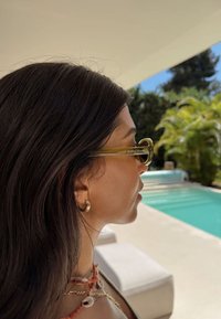 Šviesiai žali akiniai nuo saulės su apvaliais lęšiais, plonais rėmeliais ir subtiliu logotipo elementu. Asmuo turi tamsius plaukus ir dėvi auksinį lankelio formos auskarą.