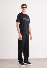T-shirt preta de algodão com o texto "BARBOUR INTERNATIONAL", combinada com calças largas pretas e ténis pretos, apresentando um design slim e óculos de sol.