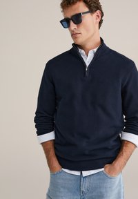 Teget pulover s teksturom i ovratnikom na patentni zatvarač, nošen preko svijetloplave košulje s gumbima. Ležeran stil s opuštenim krojem i elastičnim rubom. Naočale za sunce tamne boje.