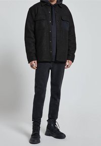Giacca camicia in misto lana nera con bottoni a pressione, due tasche sul petto, indossata sopra un felpa scura, abbinata a jeans scuri e stivali neri.