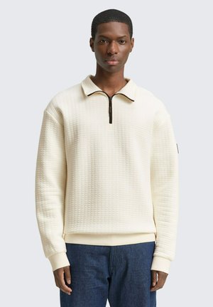 Crème-kleurige sweatshirt met een textuurpatroon, half-rits kraag en geribbelde mouwen, gecombineerd met blauwe denim broeken.